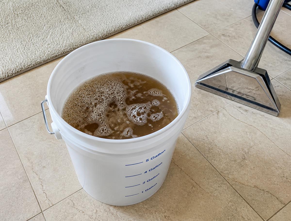 Bucket of dirty brown foam extracted from a carpet, showing old soap residue removed during cleaning